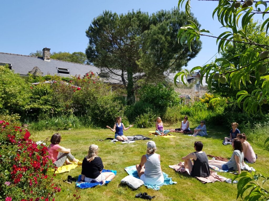 Photo globale d'une classe de yoga en extérieur, sur l'herbe, dans laquelle Gwladys est assise en tailleur face à ses élèves placés en demi cercle.